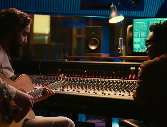 Musician and sound engineer recording guitar tunes in control room at professional music production studio. Artist playing electro acoustic instrument, live performance in soundproof space. Camera B.